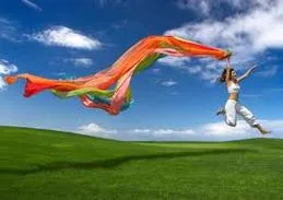 Une femme saute dans un champ d’herbe verte sous un ciel bleu, une longue écharpe de soie orange et multicolore flottant derrière elle.