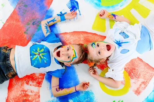 Deux jeunes enfants allongés sur une grande feuille peinte, le visage barbouillé de peinture, riant vers l’objectif.