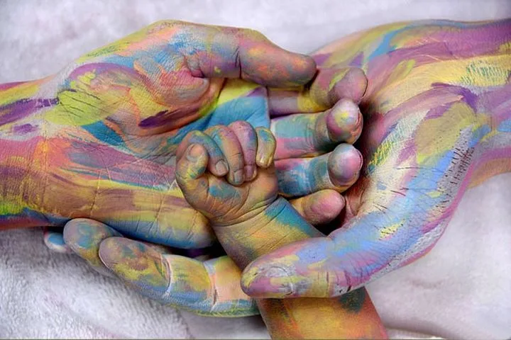 Deux mains d’adulte peintes de couleurs vives entourent la petite main d’un nouveau-né, peinte des mêmes couleurs.