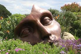 Grande sculpture de visage aux yeux clos, posée au milieu d’un jardin fleuri.